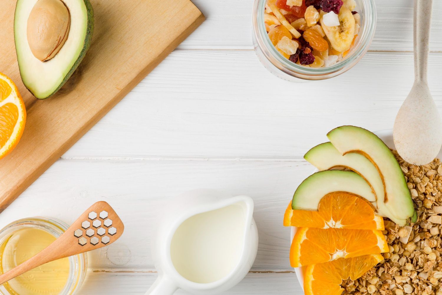 Vista desde arriba de los ingredientes del desayuno: aguacate, rodajas de naranja, granola, miel y leche sobre una superficie de madera blanca.
