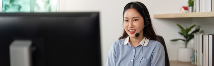 A professional wearing a headset smiles while working at a computer in an office setting with shelves and a plant.