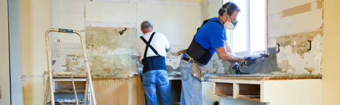 Two workers in blue shirts and work gear renovate a kitchen with exposed walls, working near a counter and a step ladder.