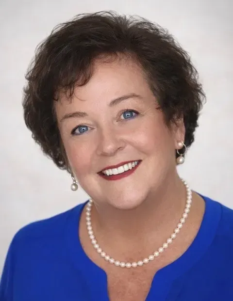 A woman in a blue shirt and pearl necklace is smiling for the camera.