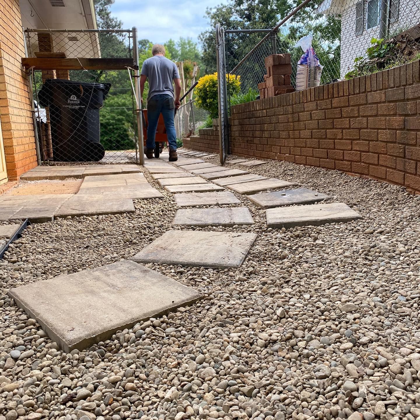 A person standing on a path of stone pavers through a gravel yard beside a brick wall and metal fence.