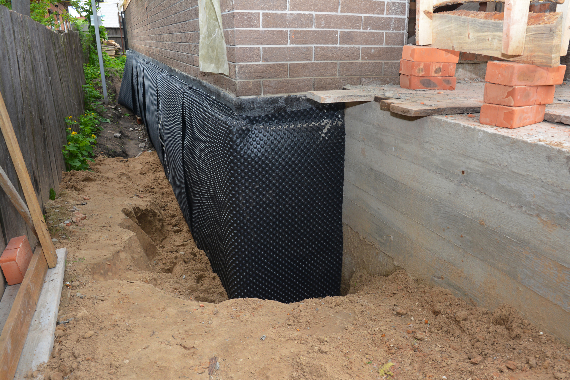Black dimpled drainage membrane installed against a house foundation wall above a dirt-filled trench.