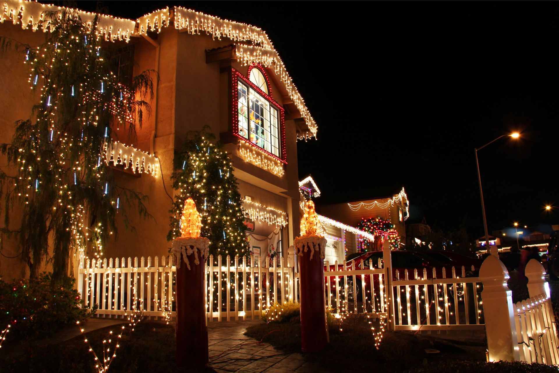 A house decorated for the holidays with bright string lights, icicle lights, and illuminated pillars behind a white fence.