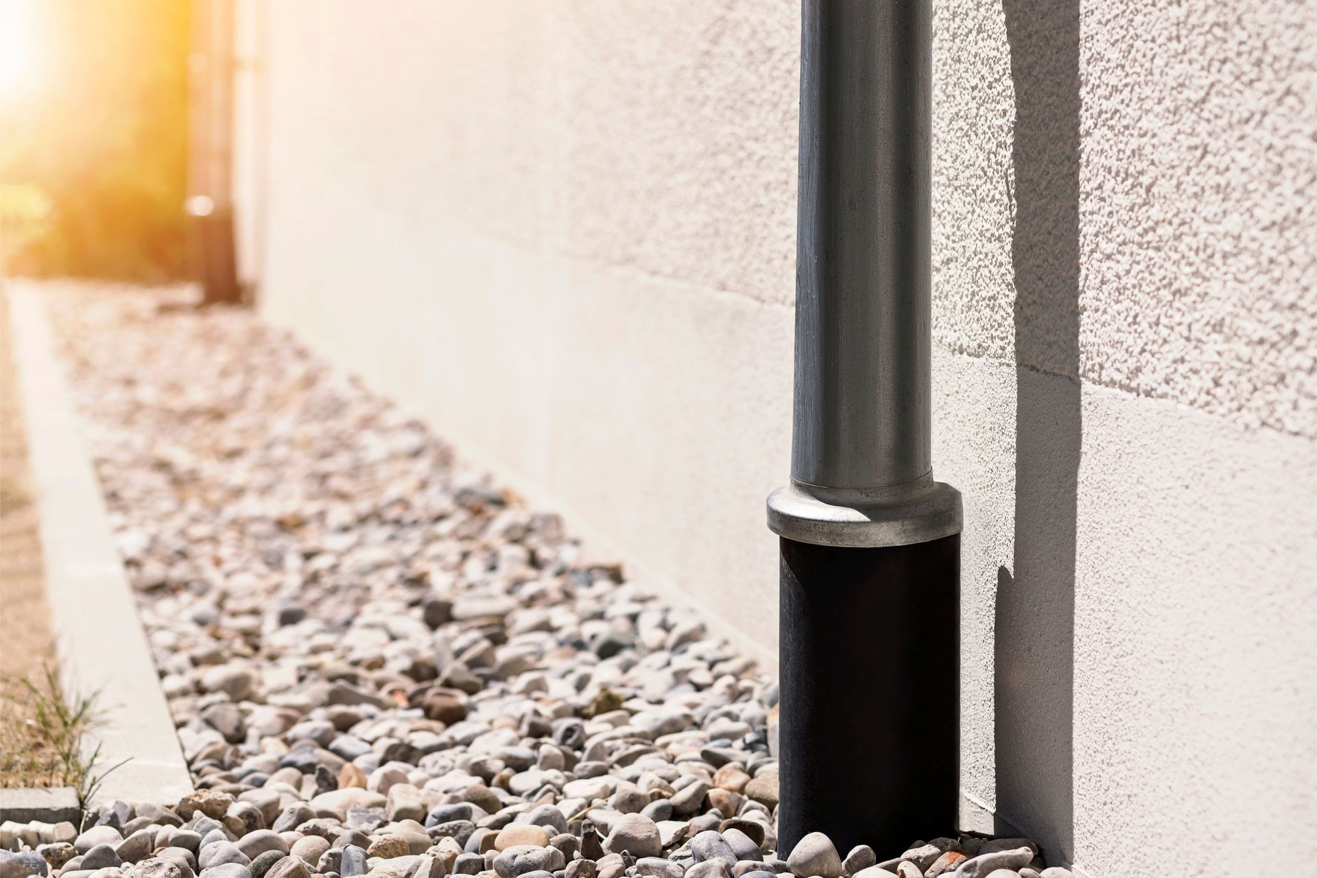 A dark gray downspout runs down a light-colored exterior wall above a gravel garden bed at sunset.
