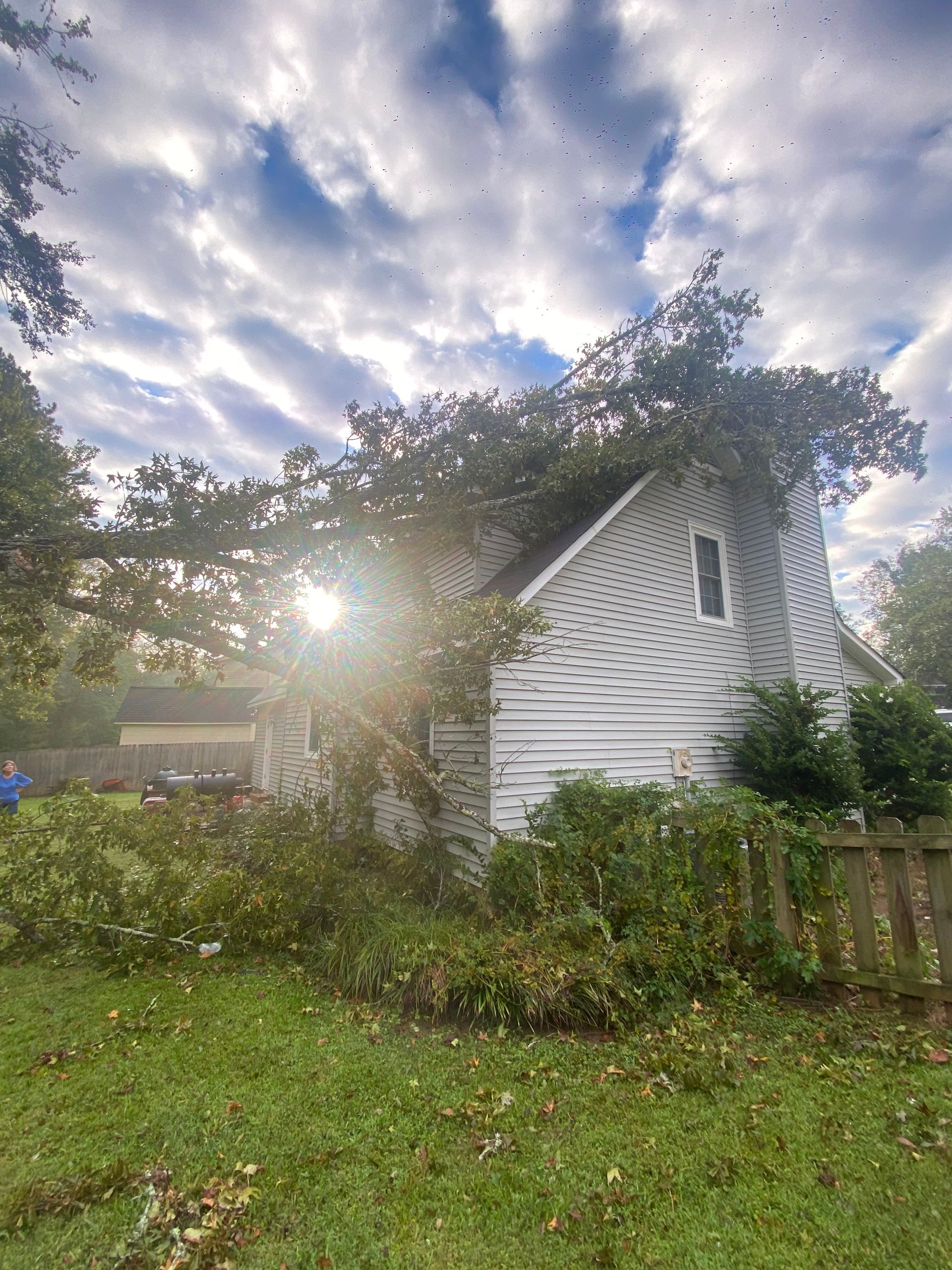 White house with slanted roof partly hidden by trees and shrubs, sunlight breaking through a cloudy sky.
