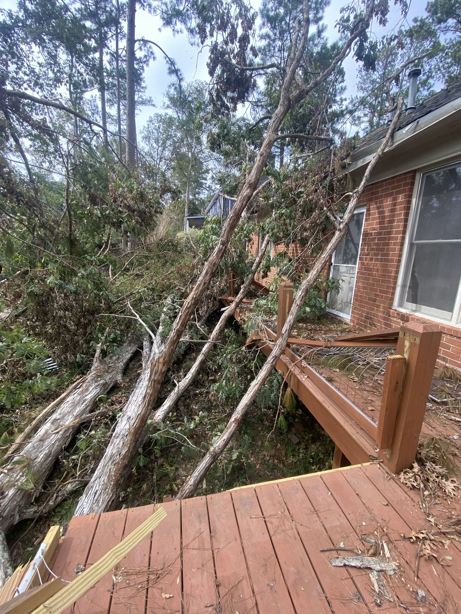 Fallen tree trunks across a wooden deck beside a brick house, with broken branches in the yard.