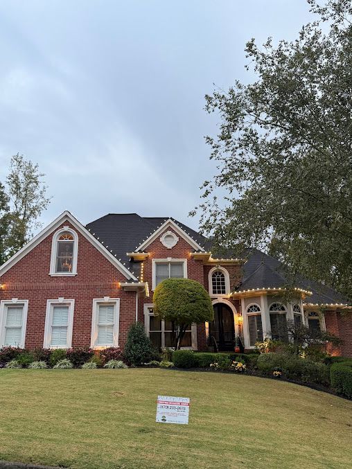 Two-story red brick house with dark roof, decorated with string lights, set behind a lawn with a small rectangular sign.