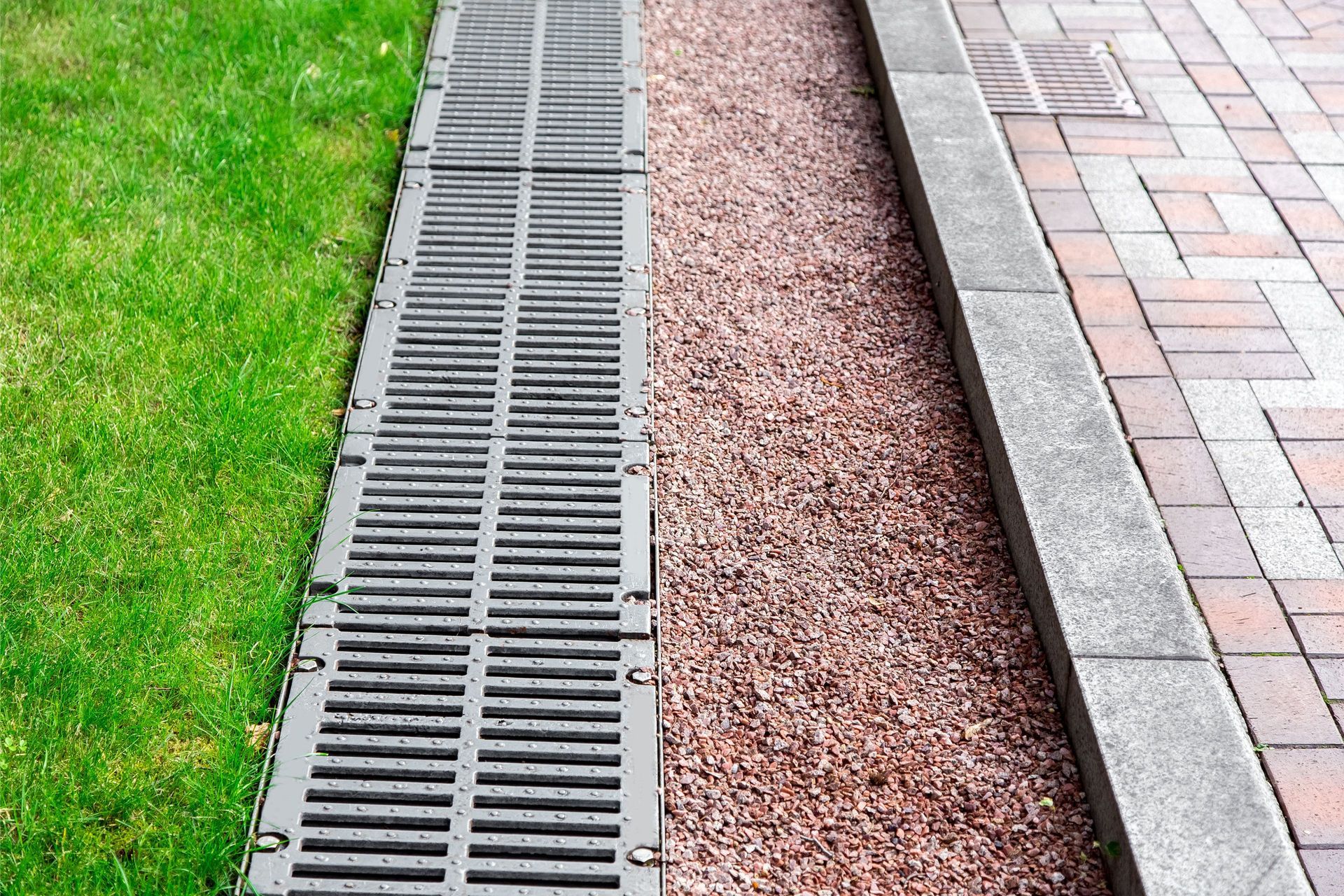 A metal drainage grate set between a grassy lawn and a red gravel strip adjacent to a paved brick sidewalk.