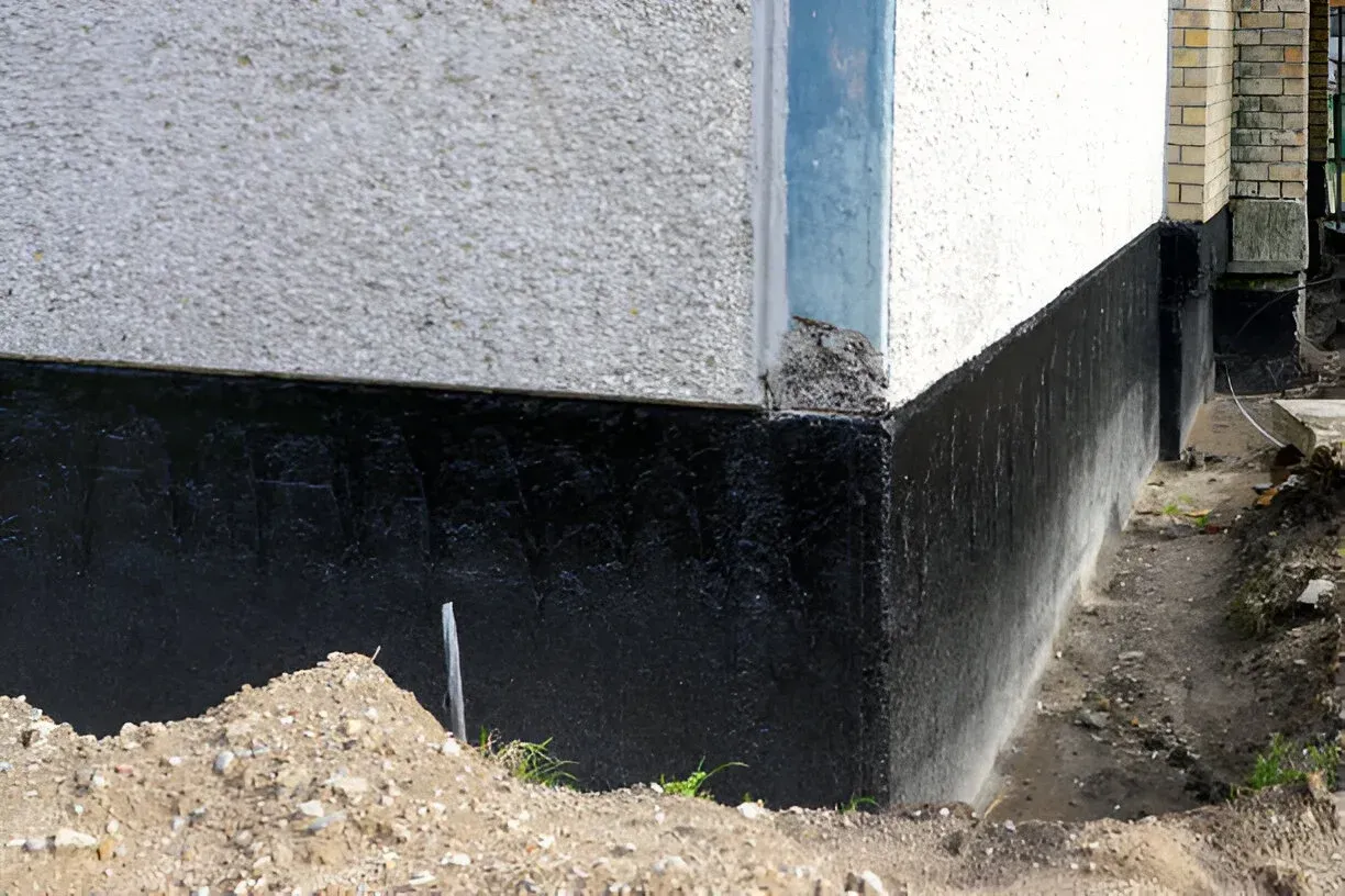 A building corner with a white stucco wall above a black, tar-coated foundation base, next to a trench of excavated soil. A building corner with a white stucco wall above a black, tar-coated foundation base, next to a trench of excavated soil.