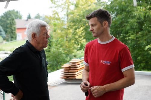 Zwei Männer stehen nebeneinander und unterhalten sich. Einer der Männer trägt ein rotes Hemd.