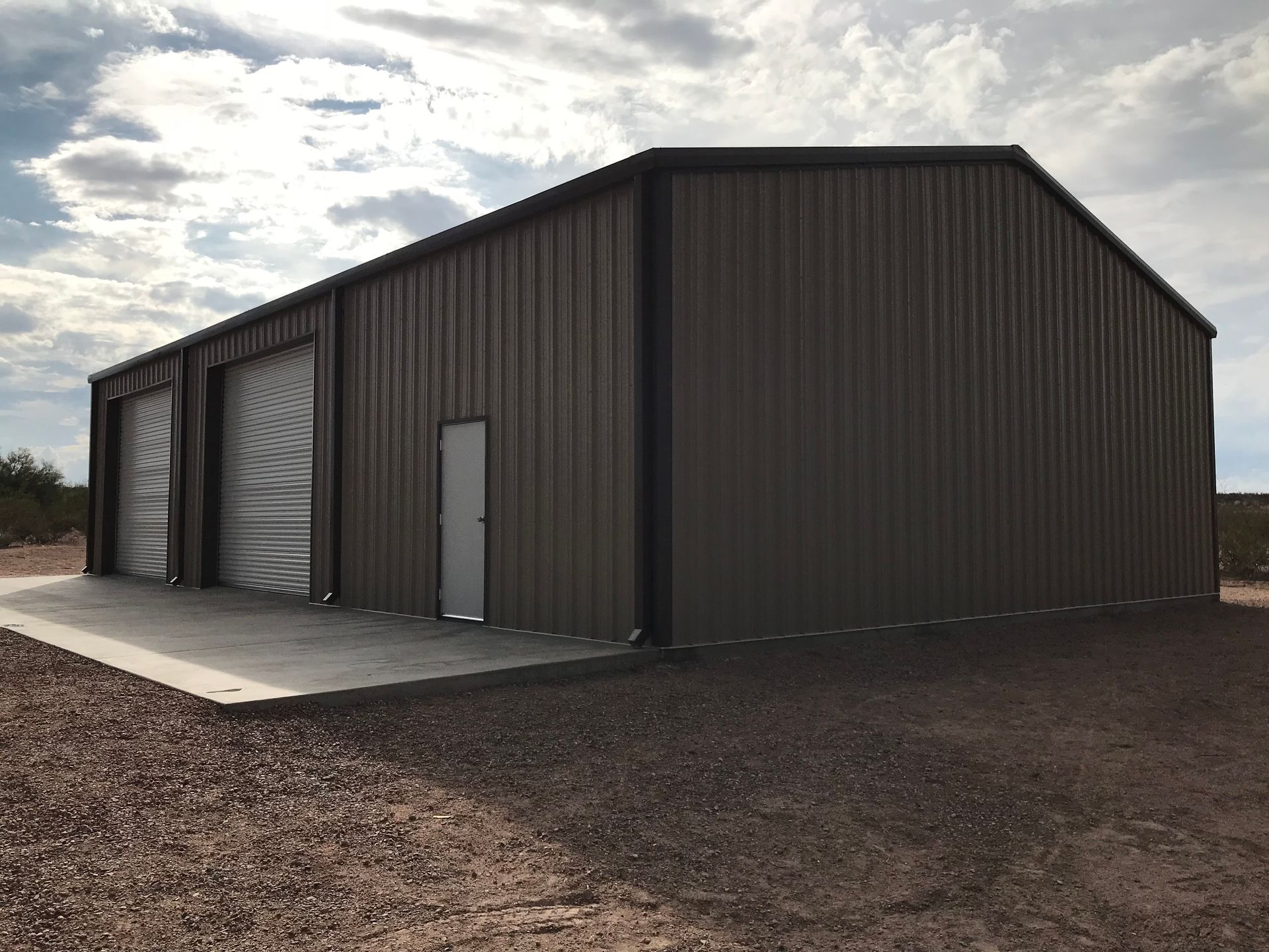 A large metal building with three garage doors is sitting in the middle of a dirt field.