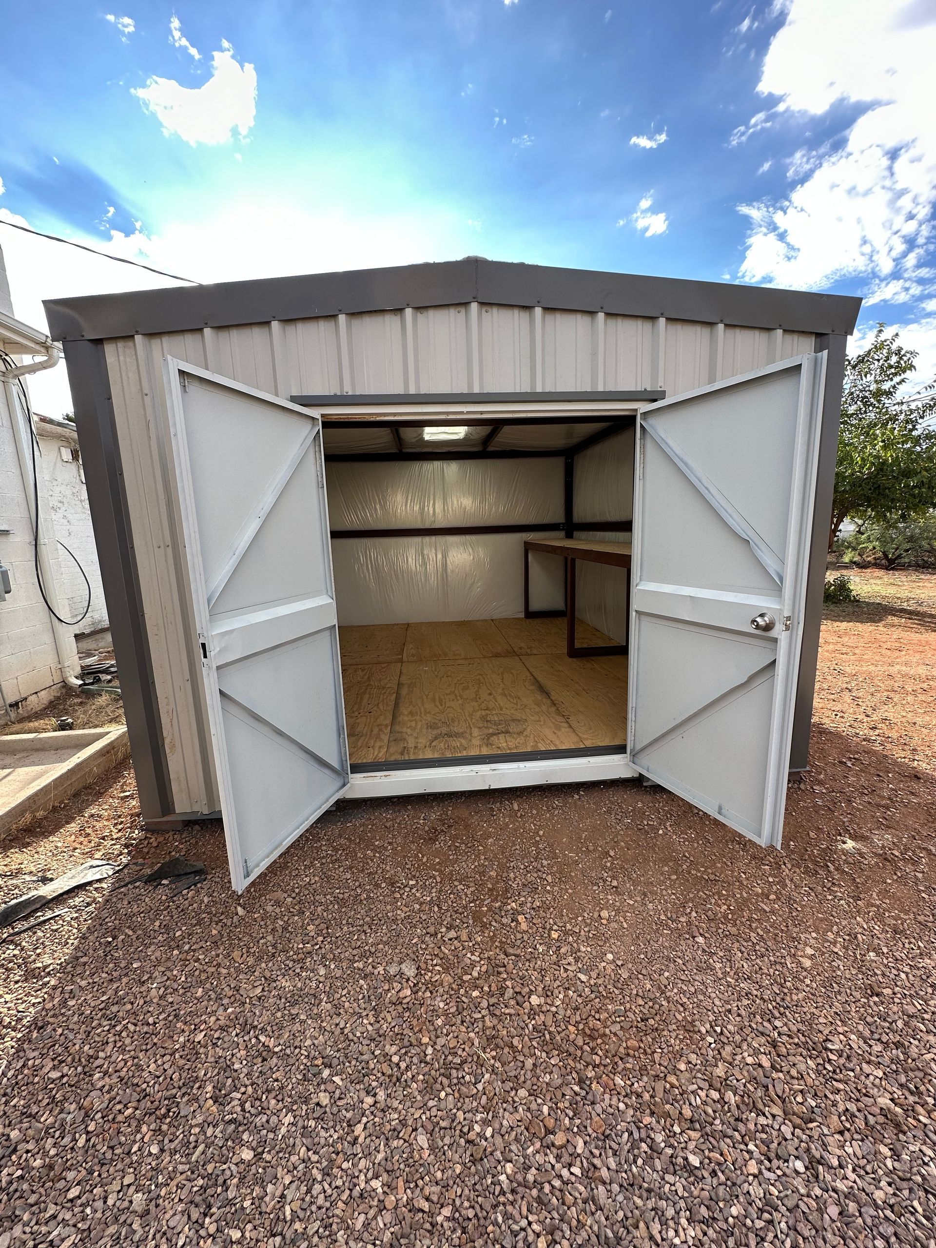 A shed with its doors open is sitting on a gravel lot.