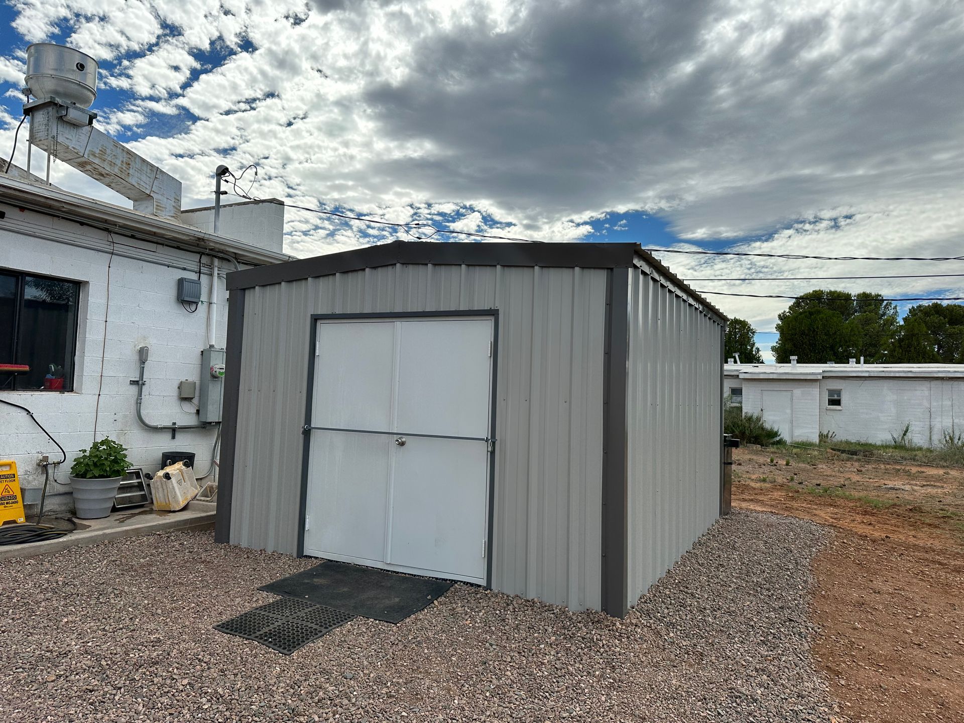 A metal shed is sitting on top of a gravel lot in front of a building.