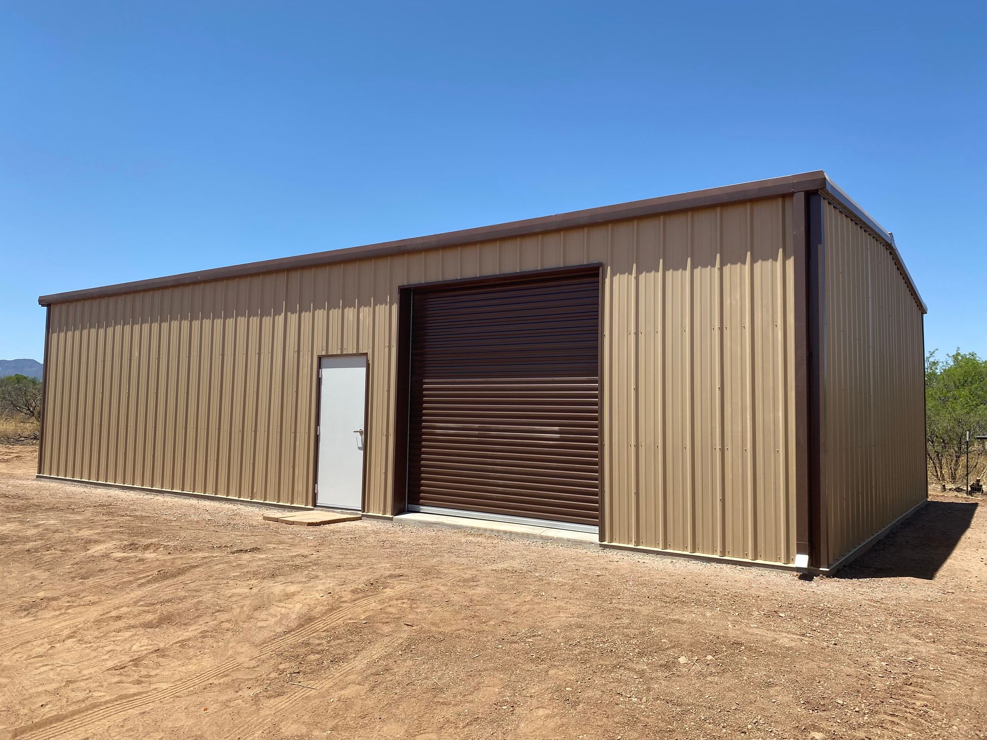 A large brown metal building with a brown garage door is sitting in the middle of a dirt field.