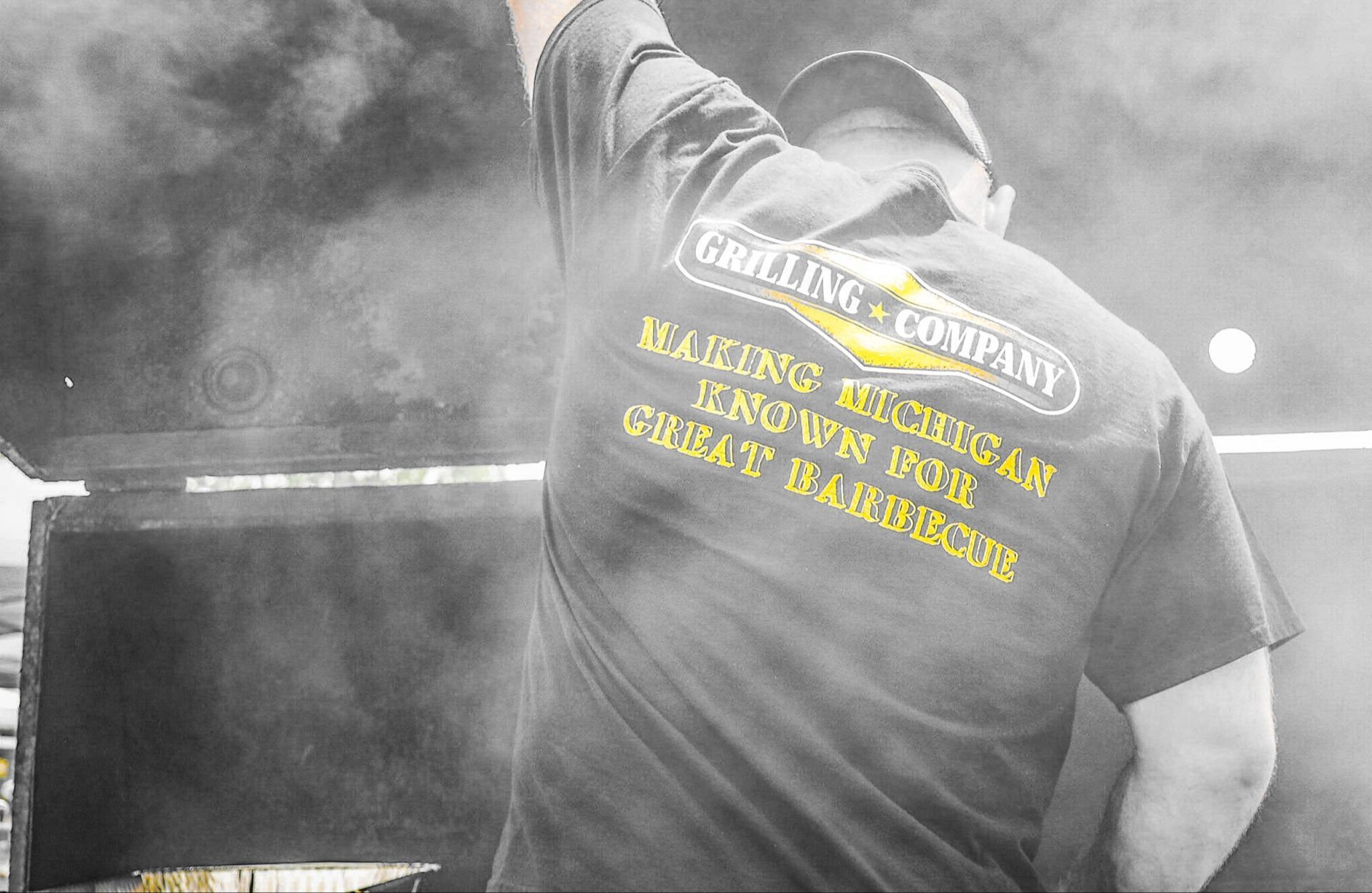 A man wearing a shirt that says grilling company is standing in front of a grill.