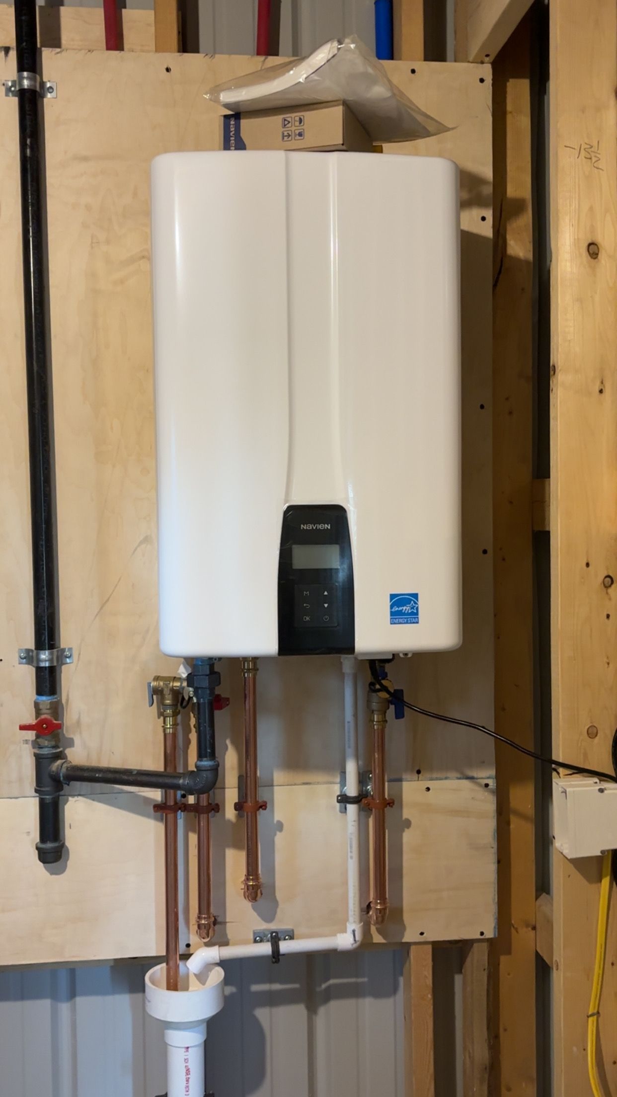 White tankless water heater mounted on plywood wall with copper pipes.