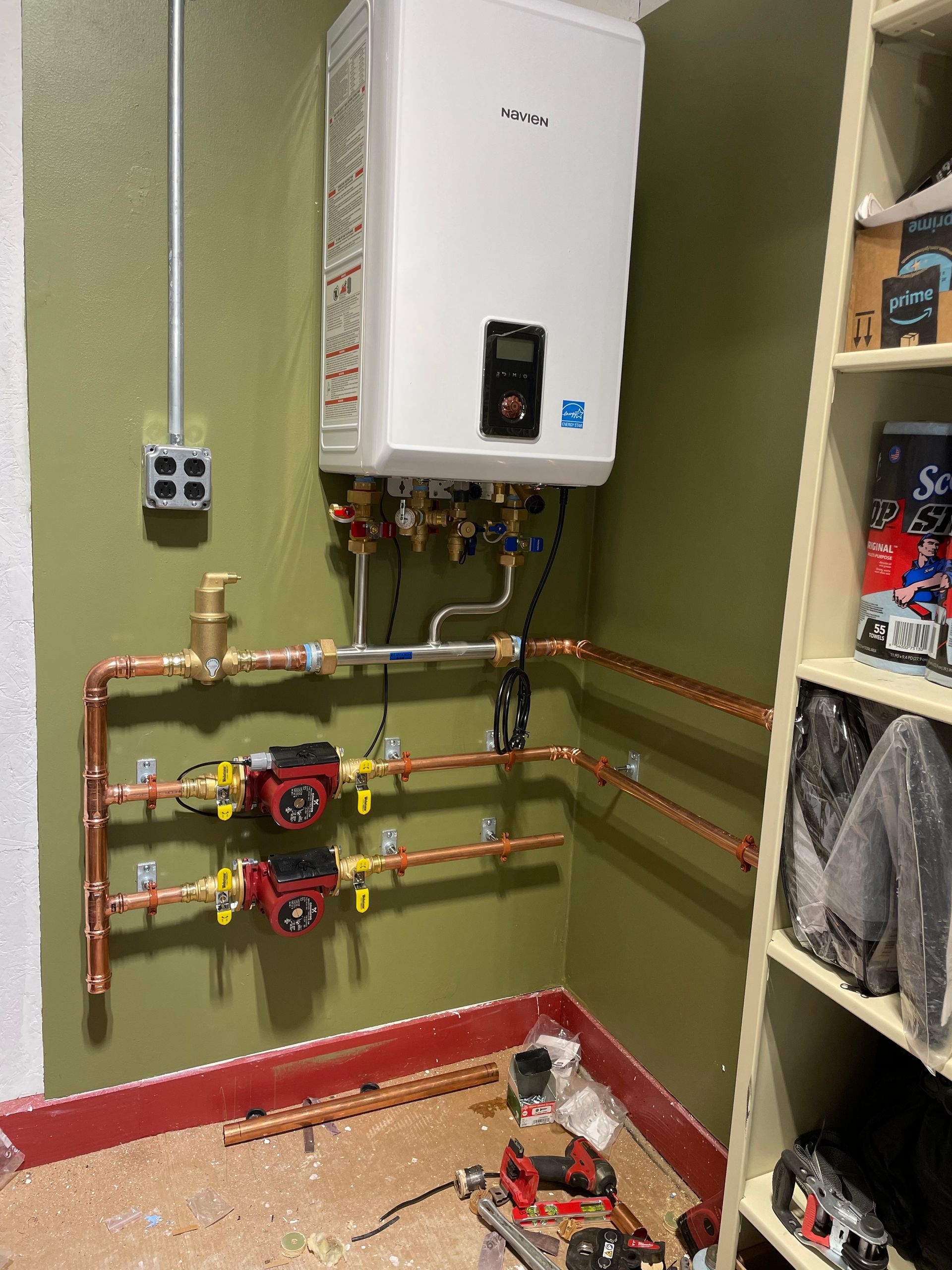 Wall-mounted water heater with copper pipes, pumps, and valves installed on a green wall in a utility space.