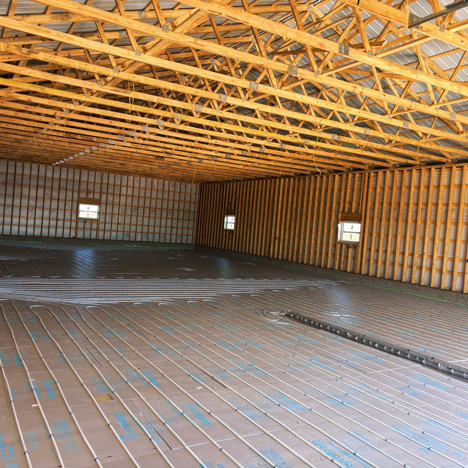 Interior of a large building under construction, featuring exposed wooden beams, concrete floor with radiant tubing, and three windows.