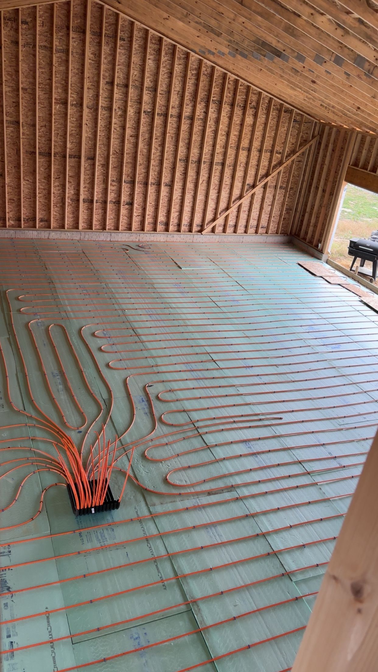 Pipes laid on a green surface for radiant floor heating in a room under construction. Orange tubes connect to a central hub.