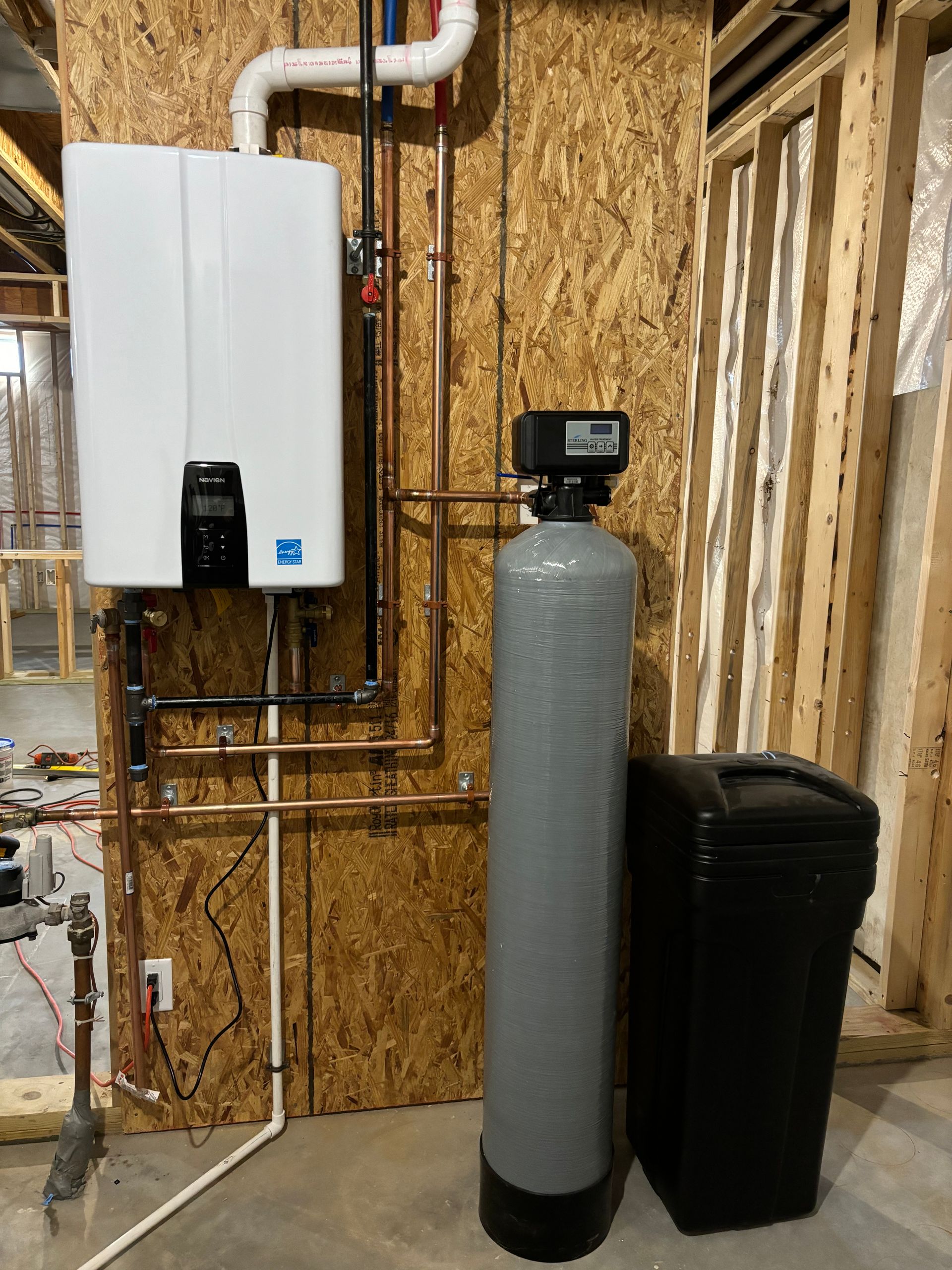 A water heater, water softener, and salt tank installed on a wooden wall. Copper pipes and wiring are also visible.