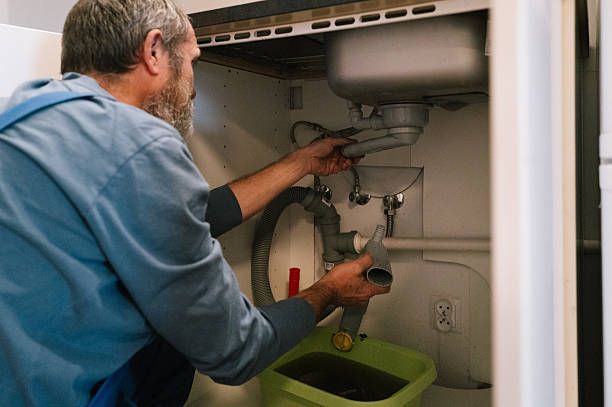 Man in blue overalls repairs sink pipes, holding a pipe section. Green bucket below collects water.