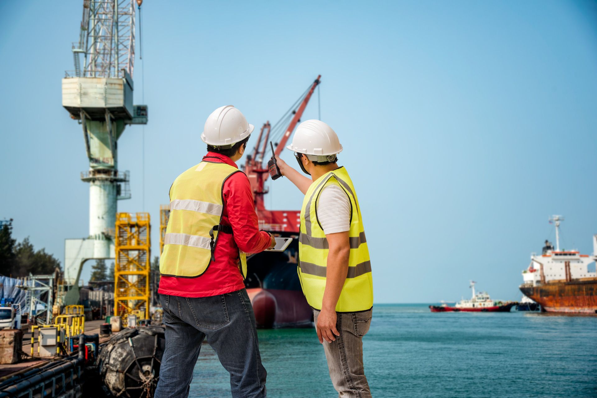 Two construction workers are standing next to each other in front of a large ship in a harbor.
