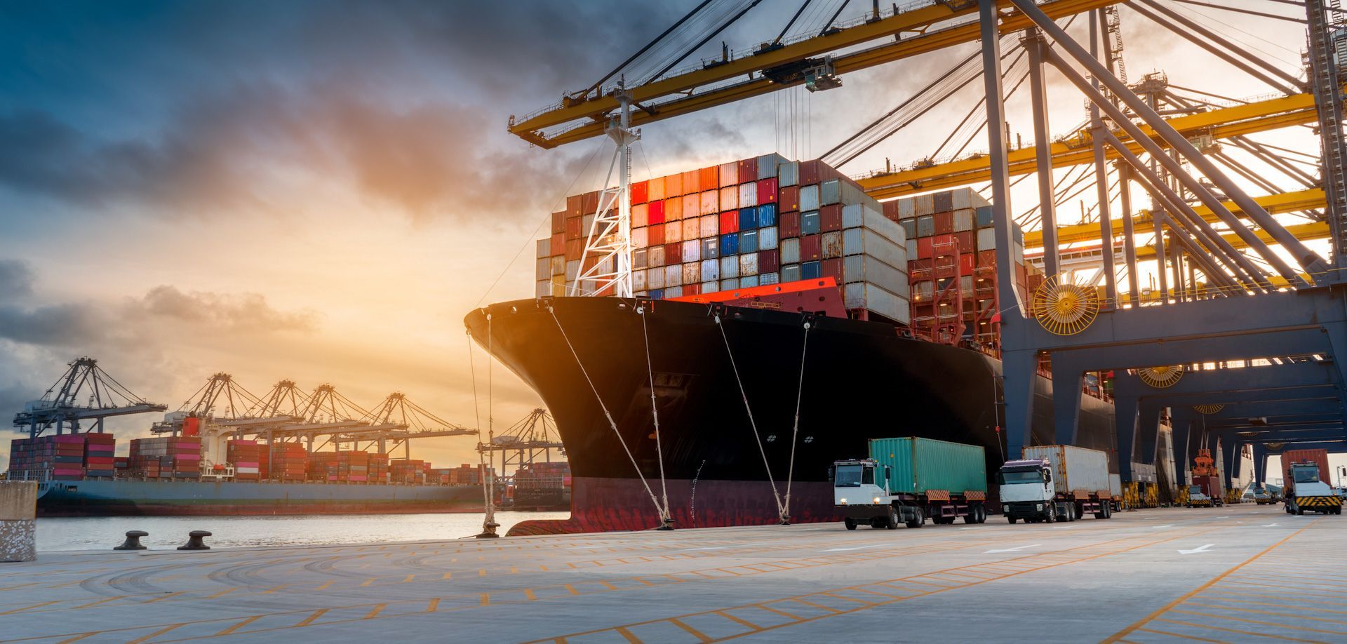 A large cargo ship being loaded with containers at a port, cranes overhead. Trucks are nearby.