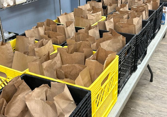 A bagged lunch including a banana, orange, sandwich, soup, eggs.