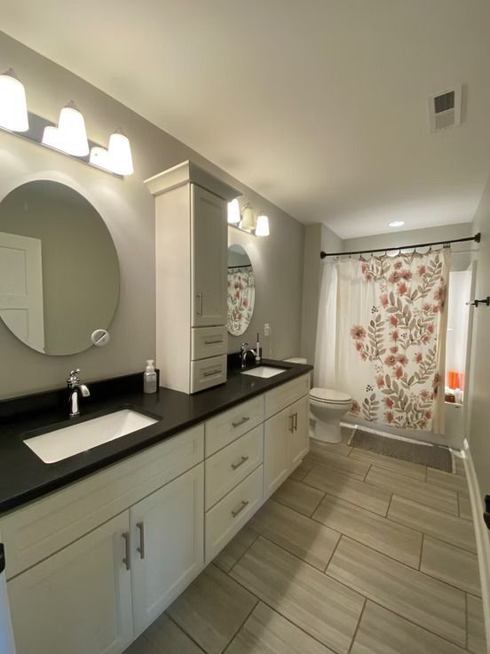 Bathroom with double vanity, round mirrors, and floral shower curtain. Light gray walls, wooden tile floors.