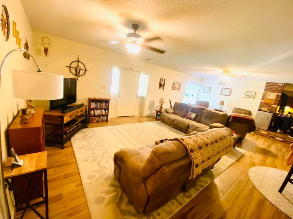 Living room with brown sectional, wooden floors, and beige rug. TV, bookshelf, and decorative items.