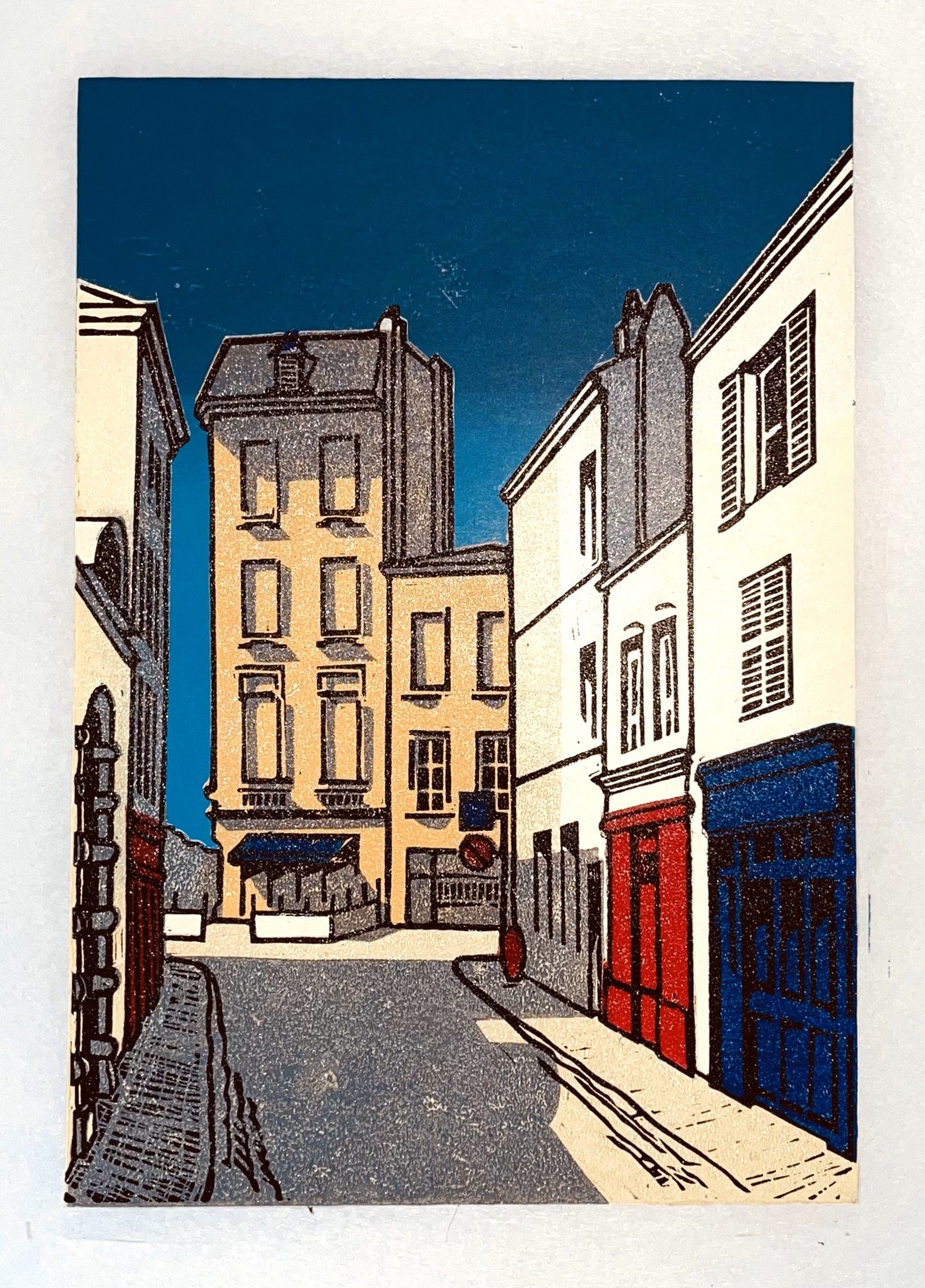 Een linosnede van een stadsstraat met gebouwen en een blauwe lucht.