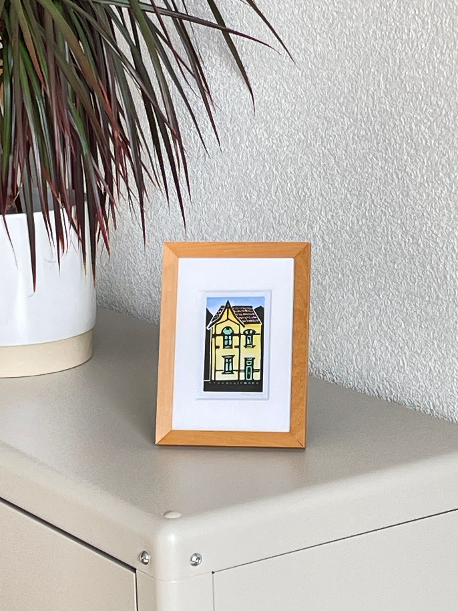 Een klein ingelijst kunstwerkje van een gebouw staat op een plank naast een plant in een witte pot.