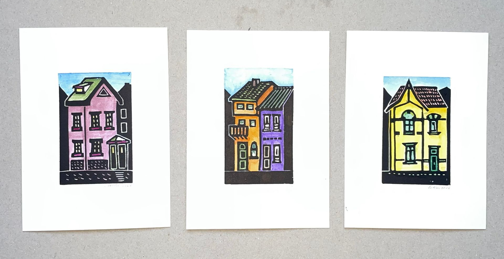 Drie kleurrijke linosnedes van huizen op een witte achtergrond, geplaatst tegen een grijze achtergrond.