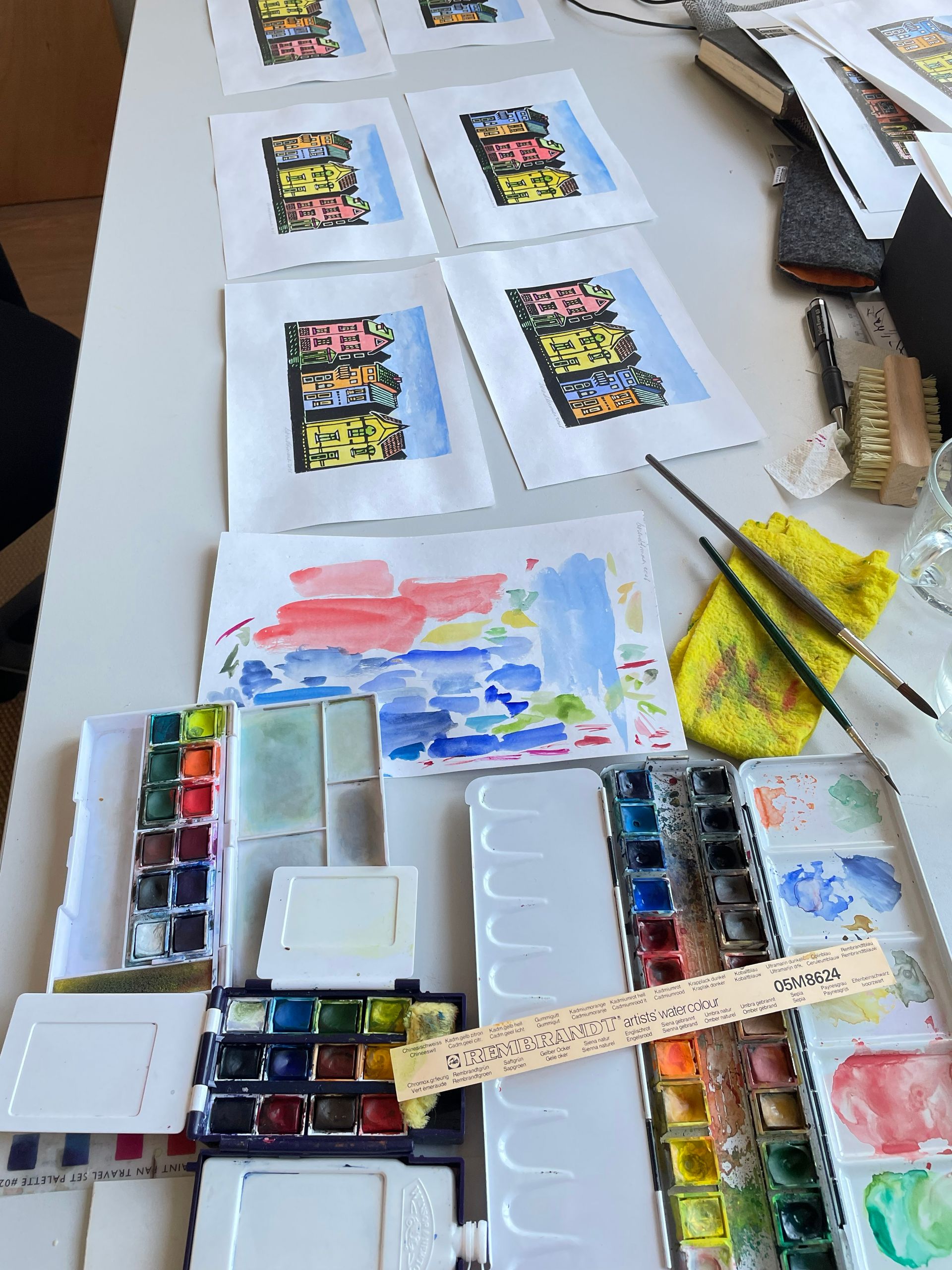 Op een wit bureau staan ​​verschillende aquarellen van gebouwen, met verfpaletten, penselen en proefpapier in de buurt.