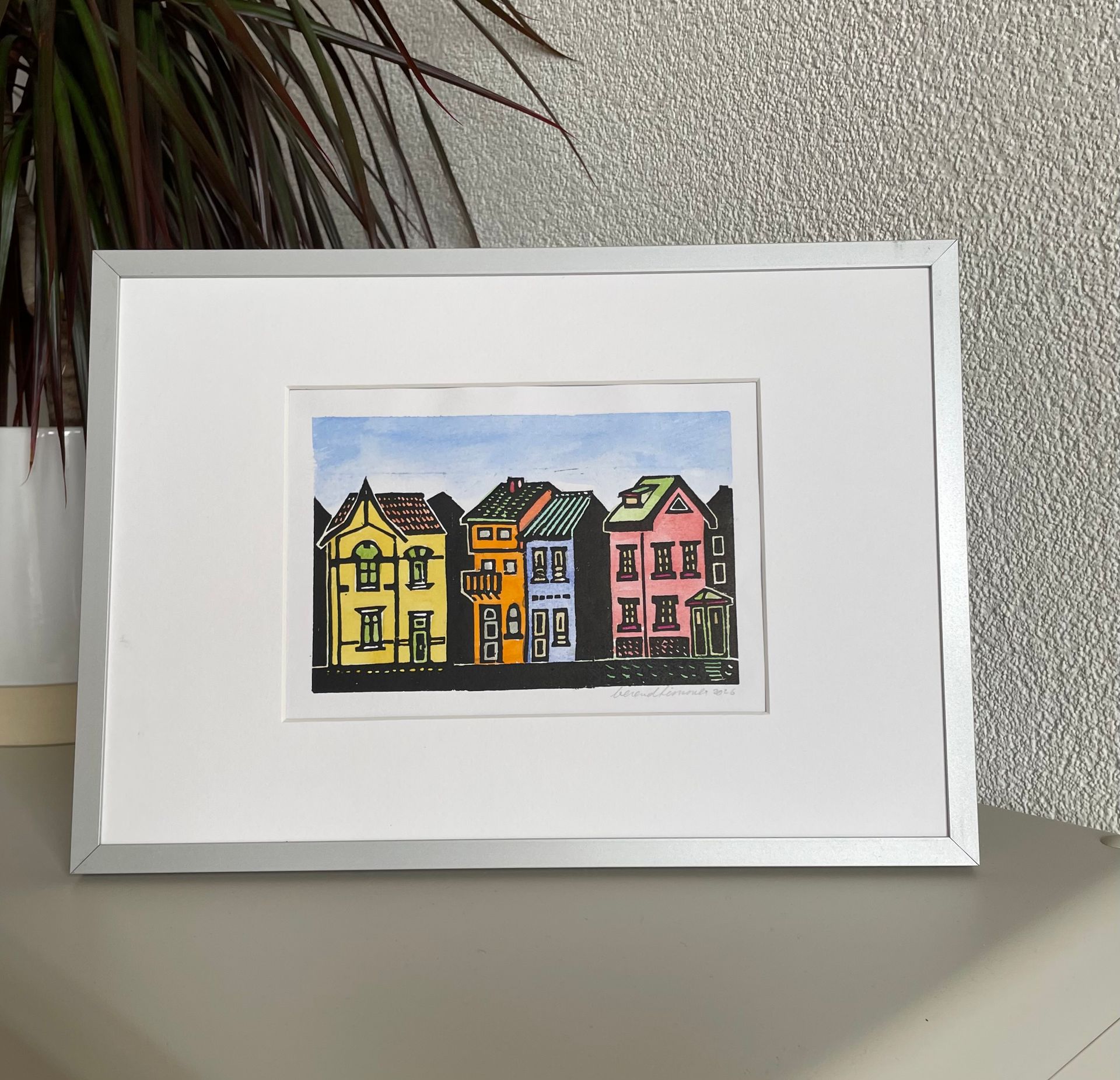 Een ingelijst aquarel schilderij van vier kleurrijke, gestileerde huizen op een rij onder een lichtblauwe hemel.
