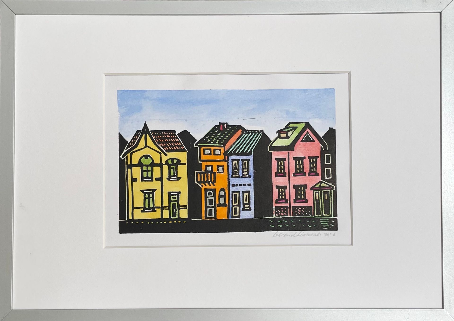 Een ingelijst aquarel schilderij van drie kleurrijke huizen – geel, oranje-blauw en roze – tegen een lichtblauwe hemel.