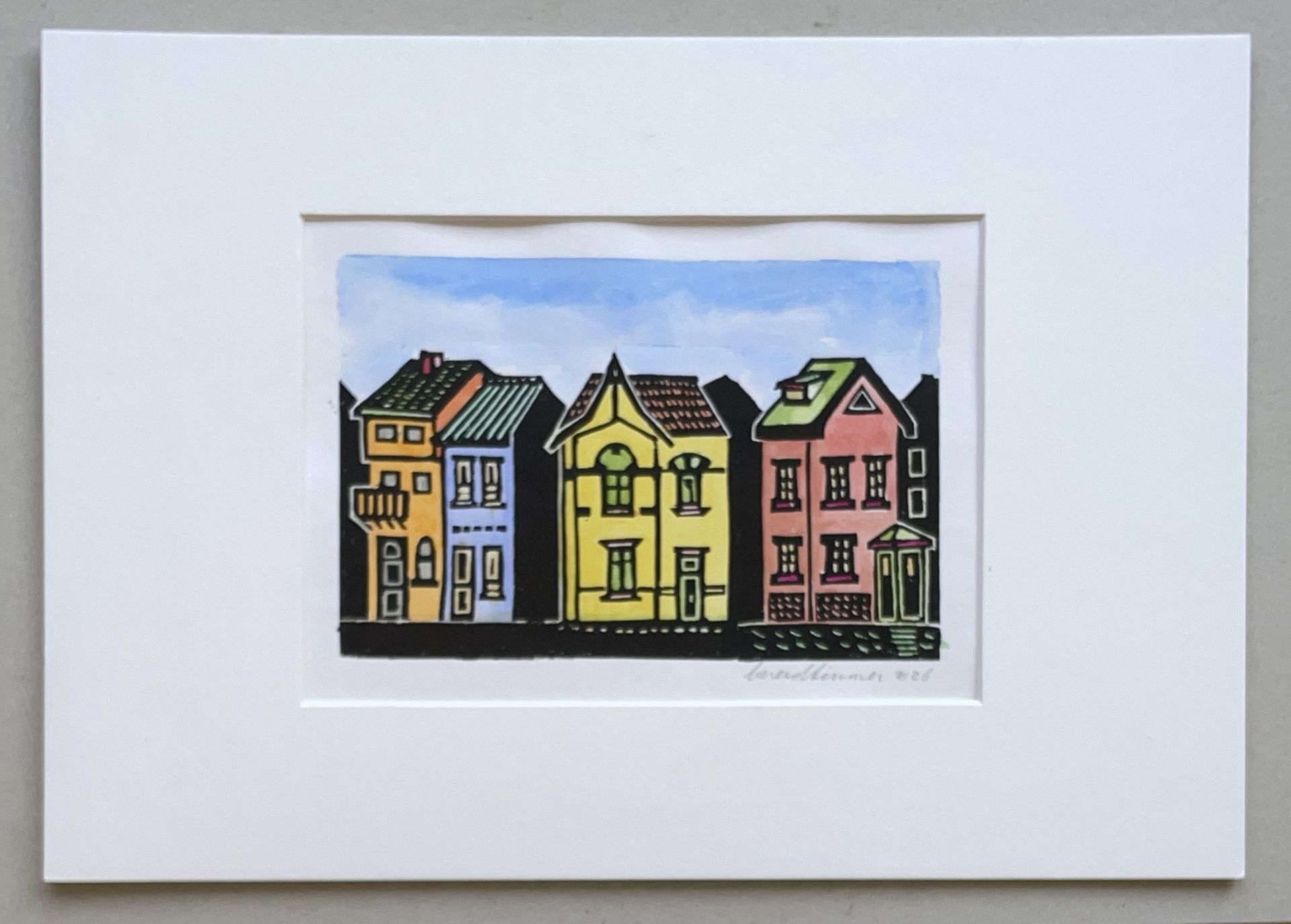 Een ingelijste linosnede van een rij van drie kleurrijke huizen in geel, roze en blauw tegen een lichtblauwe lucht.