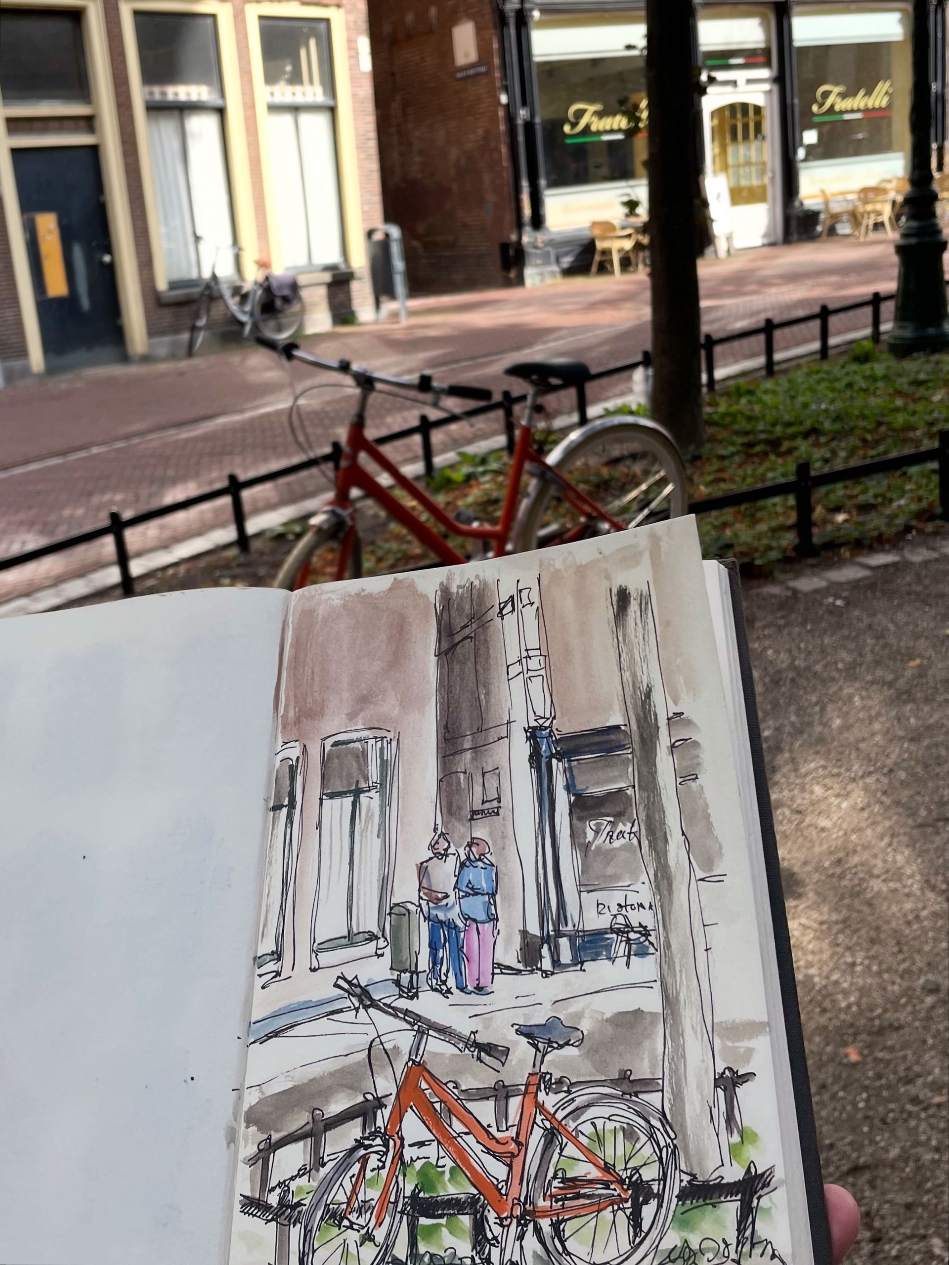 Schetsboek met een aquareltekening van een straatbeeld: rode fiets, gebouwen en mensen.