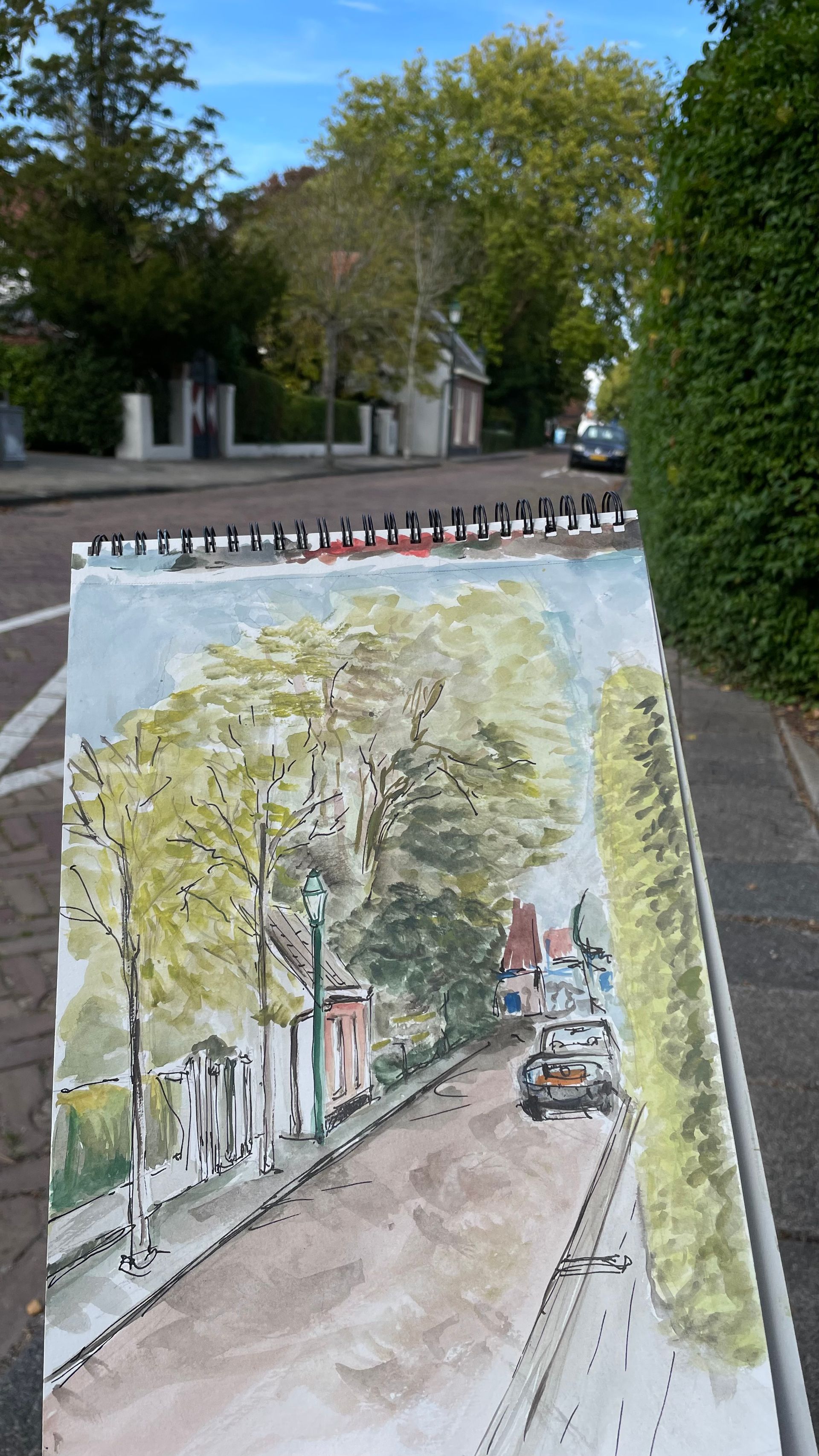 Een aquarel van een straatbeeld, met huizen, bomen en een auto, en een notitieblok op de voorgrond.