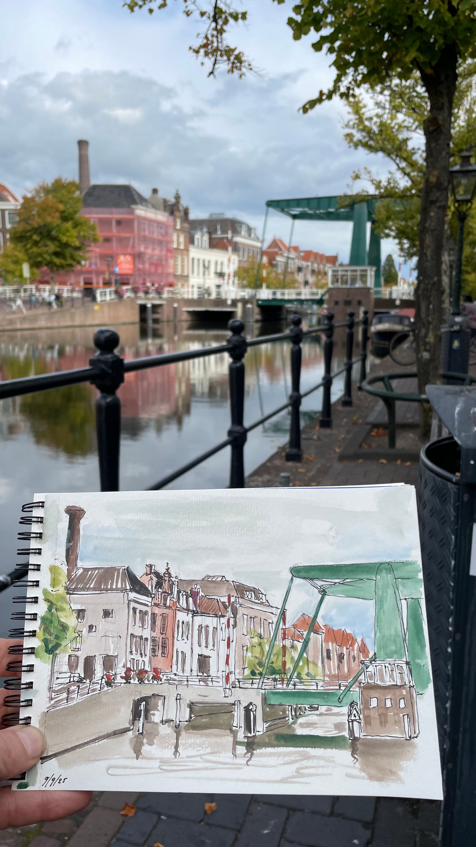 Een schets van een kanaalscène, met een groene brug, die voor het daadwerkelijke uitzicht wordt gehouden. Gebouwen en een bewolkte lucht.