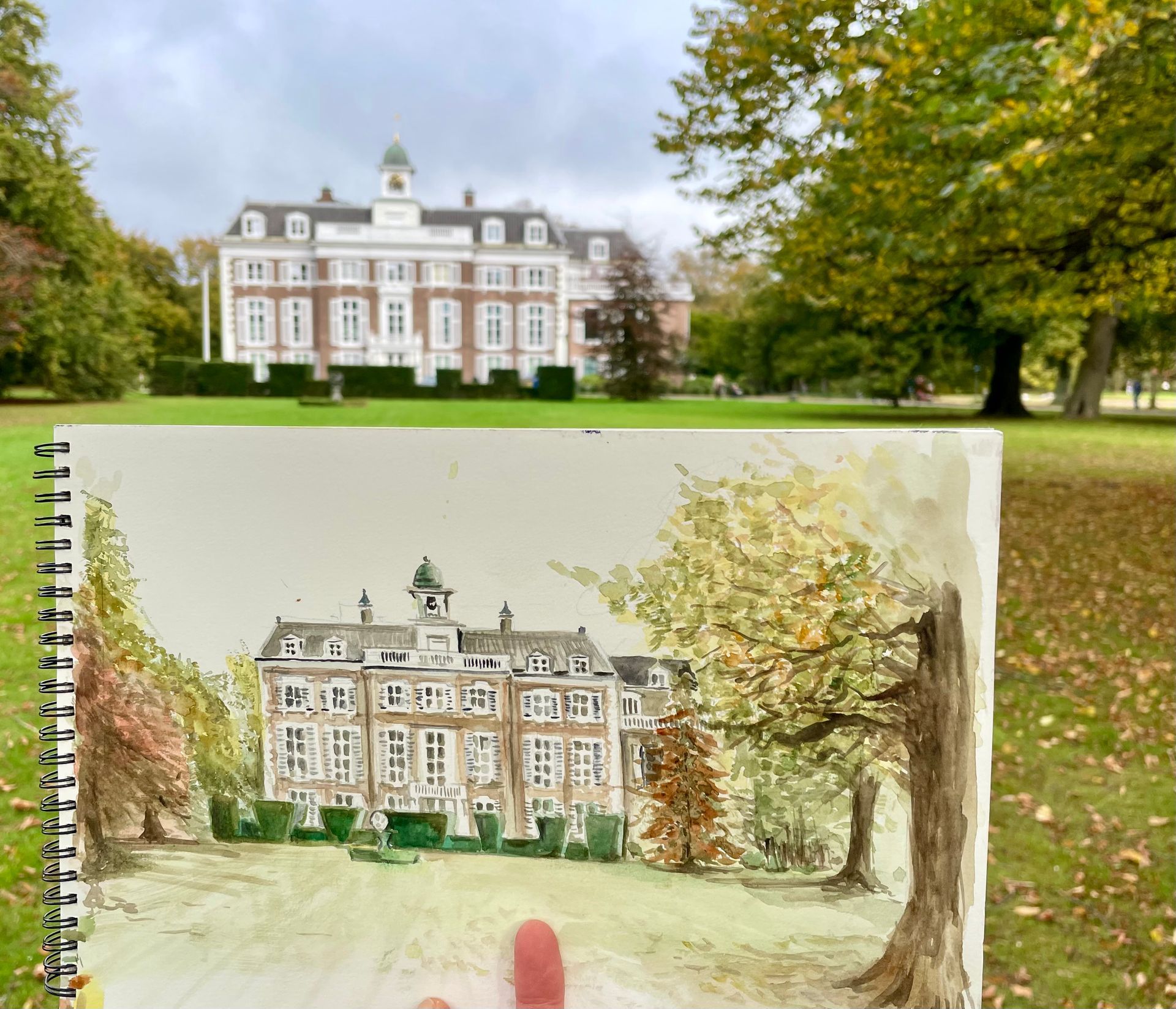 Aquarel van een statig bakstenen gebouw in een park, ter vergelijking voor het daadwerkelijke gebouw gehouden.