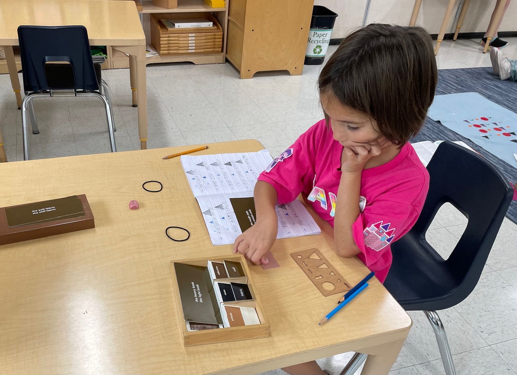A little girl in a pink shirt is sitting at a table in a classroom.