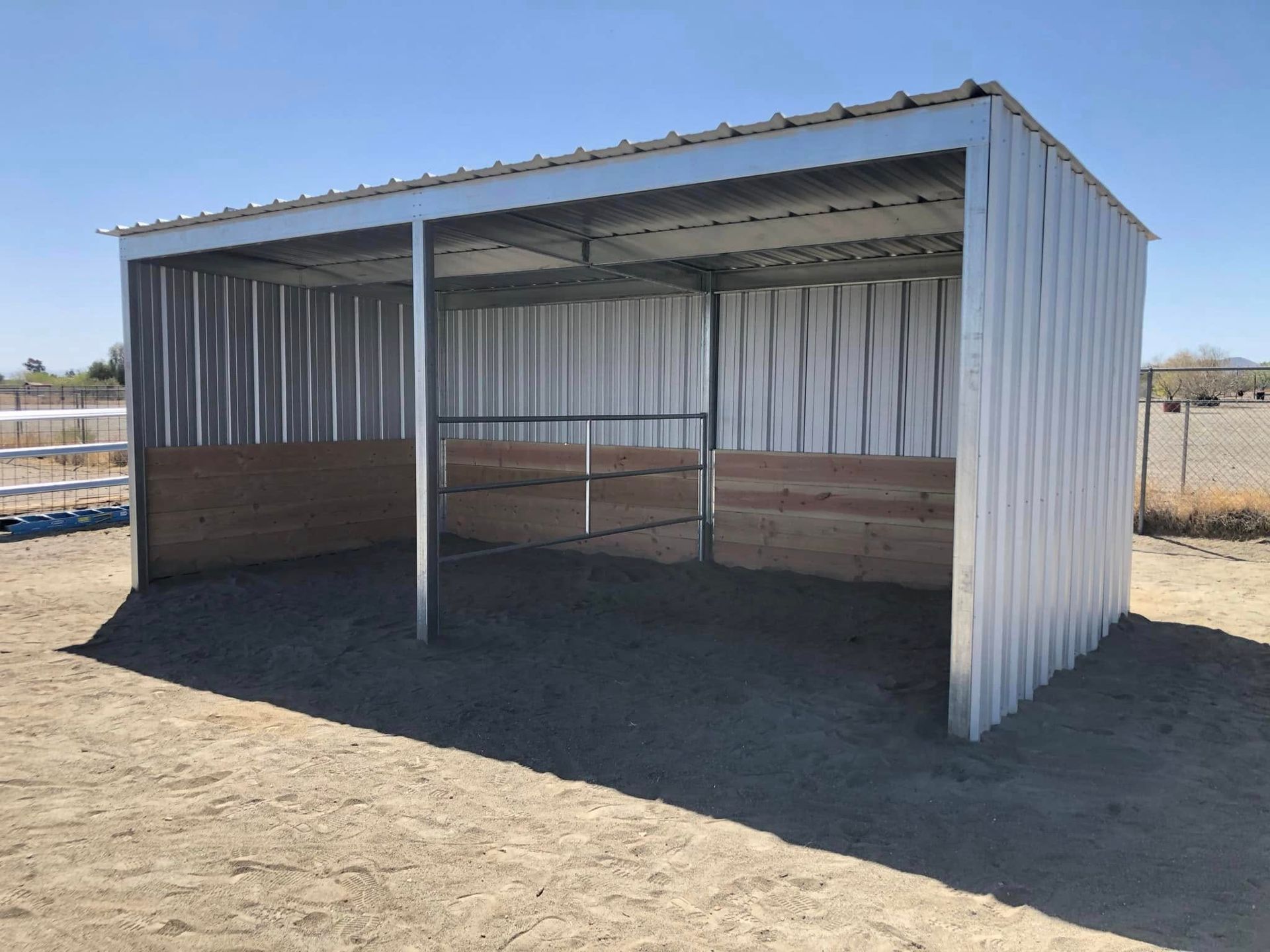 A white metal shed with wooden walls is sitting in the middle of a dirt field.