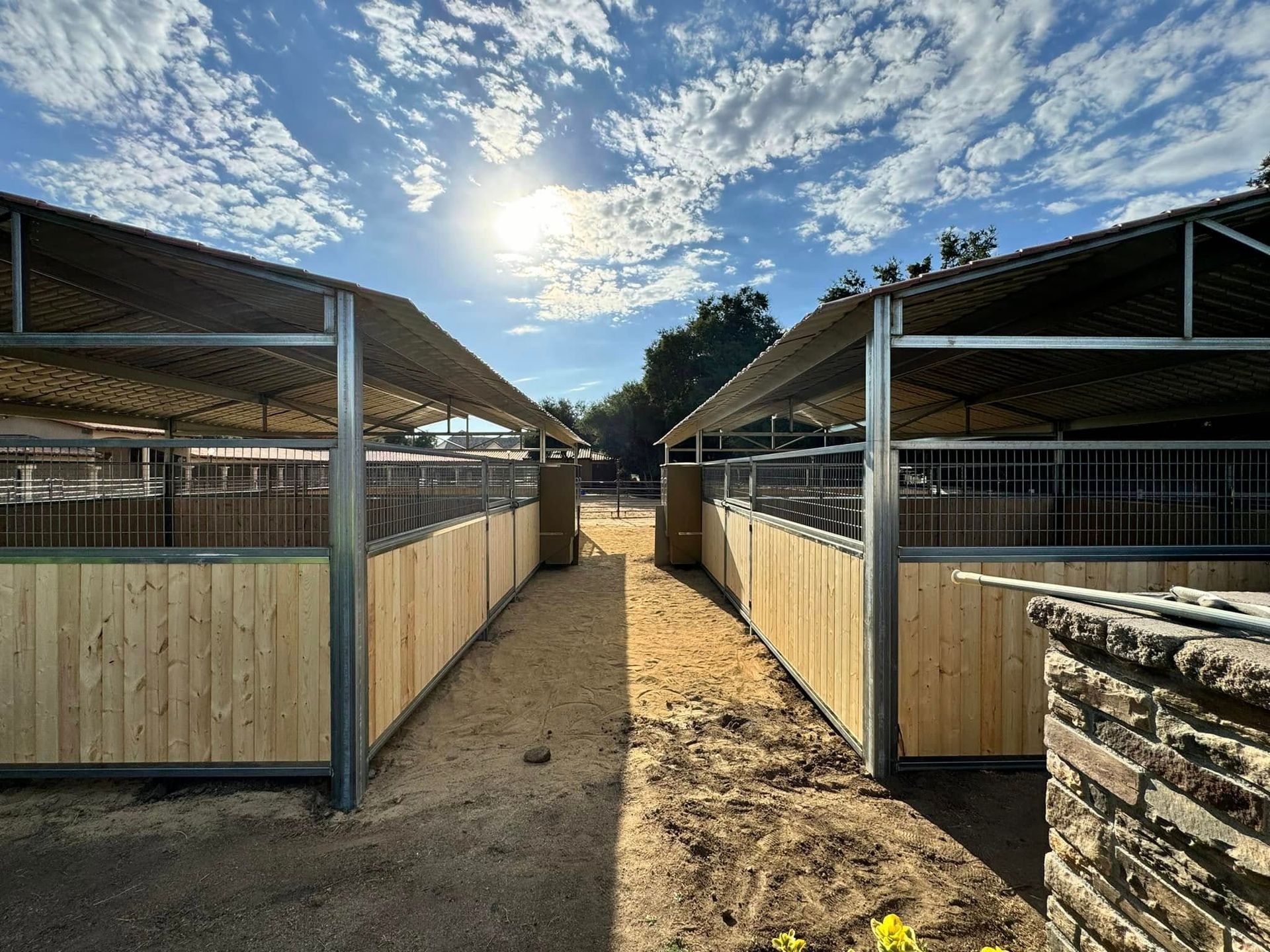 A row of wooden stables with a roof on a dirt field.