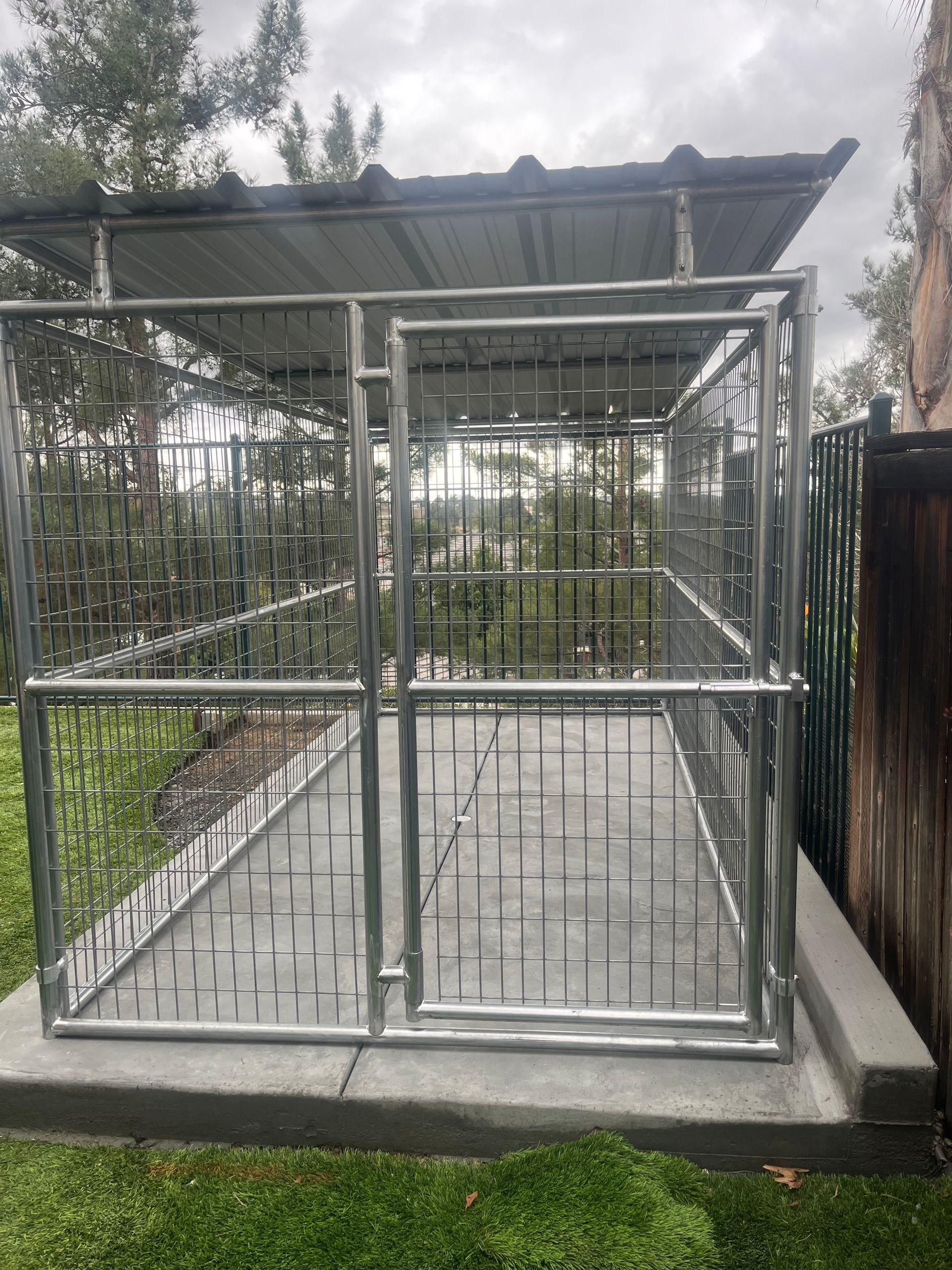 A metal dog kennel is sitting on top of a concrete platform in a backyard.
