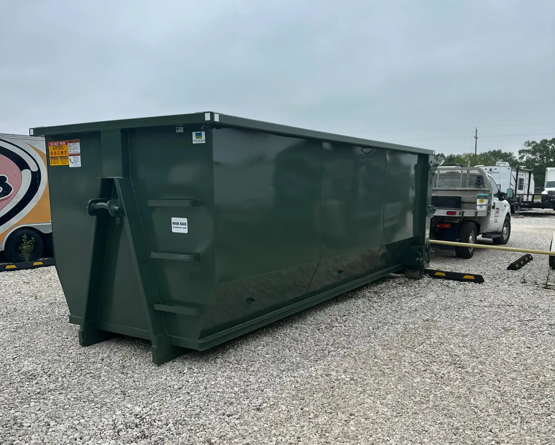 A large green dumpster is sitting on top of a gravel lot next to a truck.