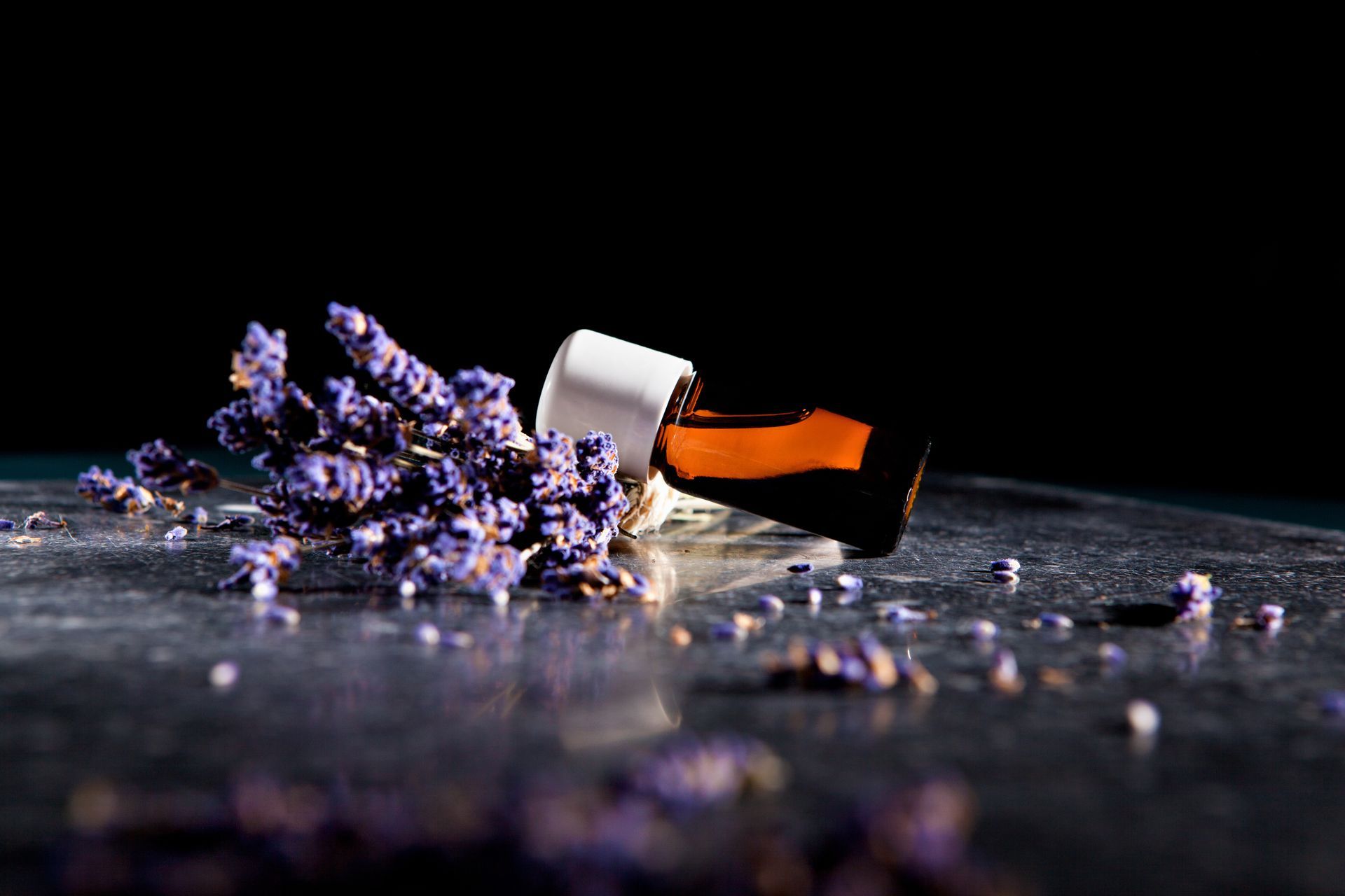 Ein kleiner, kippbarer Behälter ergießt orangefarbene Flüssigkeit und violette Kristalle auf eine dunkle Oberfläche.