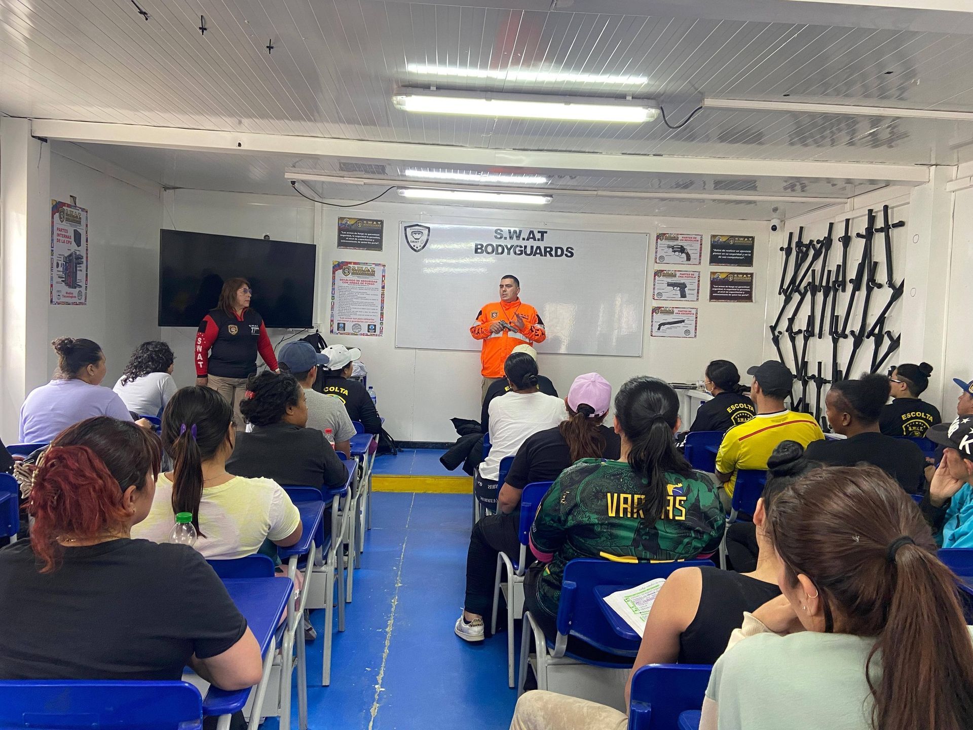 Personas en un aula; un instructor con traje naranja da una presentación. Los asistentes están sentados en sus escritorios.