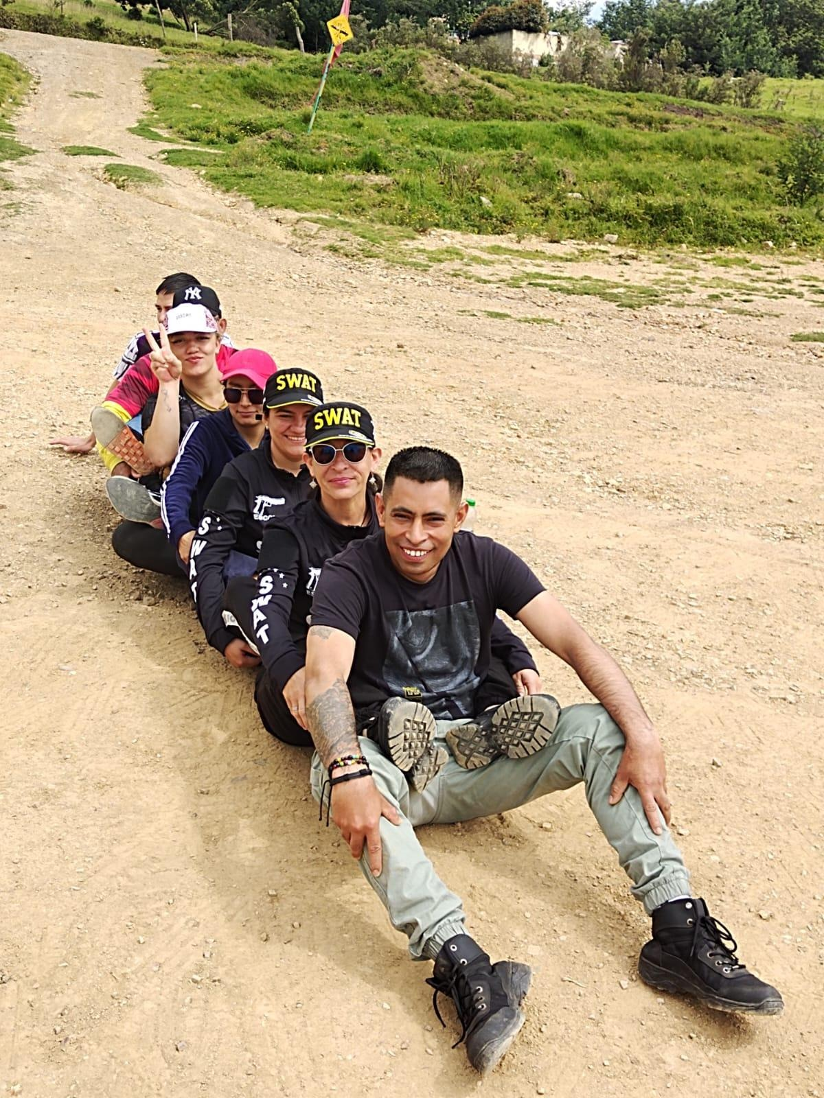 Seis personas sentadas en fila en un camino de tierra, sonriendo. Llevan sombreros y ropa informal. Al fondo, una colina cubierta de hierba.
