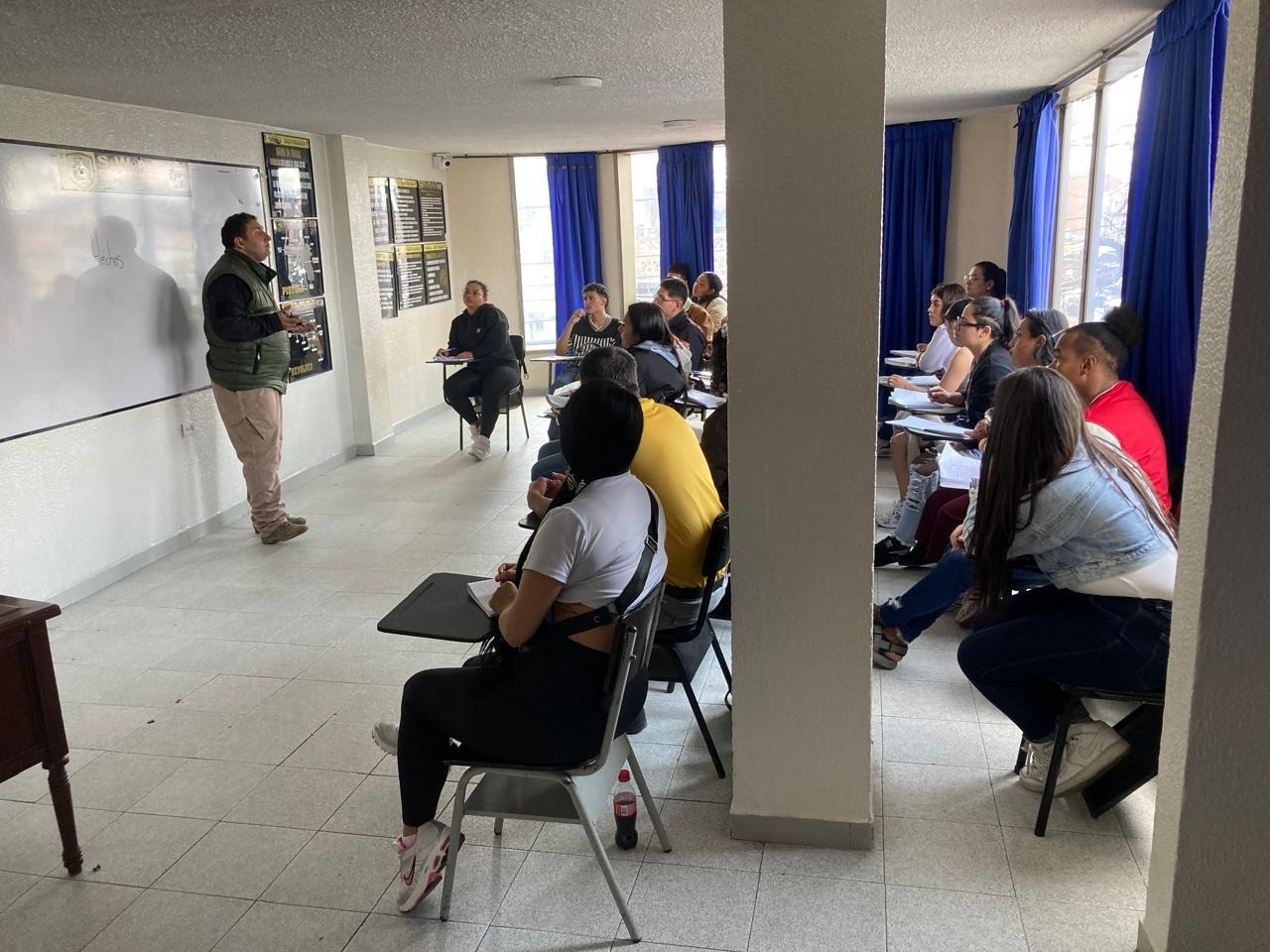 Una clase magistral: el profesor en la pizarra, los alumnos sentados. Cortinas azules, paredes claras.